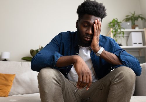 Overwhelmed African American man sitting on the couch, having anxiety and feeling sad.